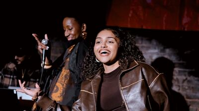 A smiling young woman in a brown leather jacket performs on a dimly lit stage while a woman behind her holds a microphone.