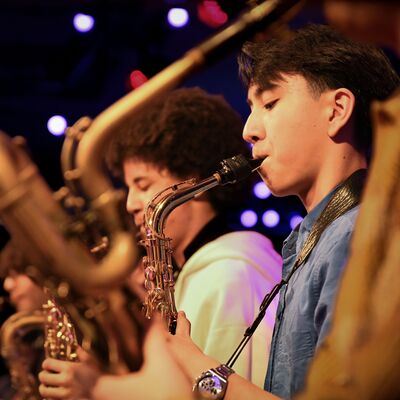 Close-up of a young man playing an alto saxophone in a line of musicians under purple and red stage lights.