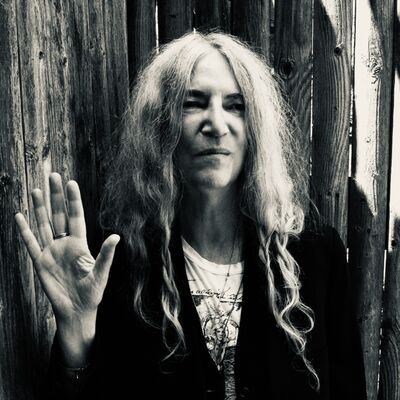 A black and white portrait of Patti Smith raising her hand with palm facing out, leaning against a wooden fence.