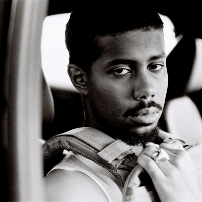 A black and white close-up portrait of a man looking over his shoulder while seated inside a vehicle, gripping a backpack strap.