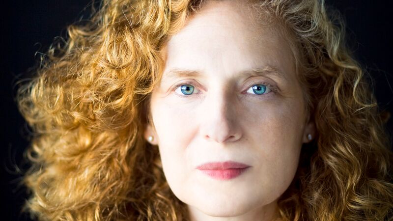 A close-up portrait of a woman with curly strawberry blonde hair and blue eyes, looking directly at the camera against a dark background.