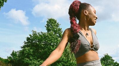 A person with long red hair in a ponytail and gold hoop earrings is dancing in front of a tall green tree