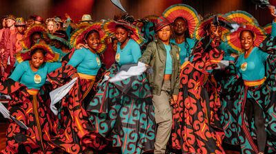 Dancers in teal tops and patterned skirts perform with colorful pleated hats on a stage lit by warm red spotlights.