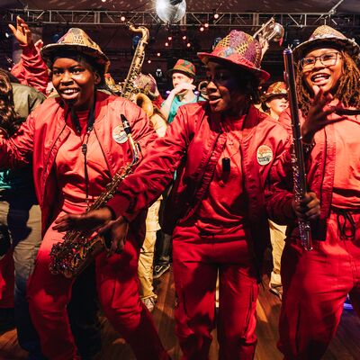 Musicians in matching red outfits and patterned hats smile while playing saxophones and flutes on a crowded dance floor.