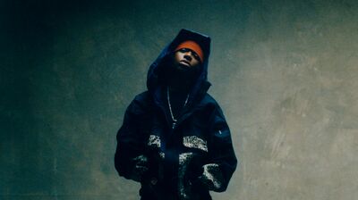 A person stands wearing baggy dark pants, a hooded jacket, and an orange beanie against a textured studio backdrop.