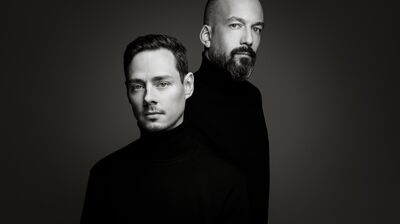 A black-and-white studio portrait of two men in black turtlenecks standing staggered against a grey gradient background.
