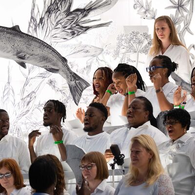 A diverse choir in white clothing sings passionately in front of a giant black and white sketch of a fish and flora.