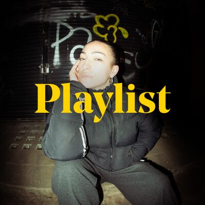 India Shan, wearing black trousers and puffer jacket sitting on a concrete step in front of a graffitied wall. The word 'Playlist' is placed over the image.