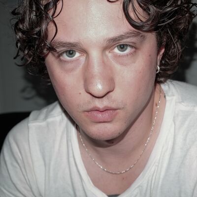 A person with brown curly hair looks into the camera wearing a white tshirt.