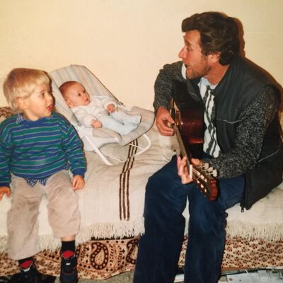 A man plays guitar on a bed for a toddler sitting beside him and a baby in a bouncer chair.