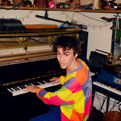 A young man in a brightly coloured sweater turns to face the camera. He is playing a piano, and is surrounded by a variety of other musical instruments