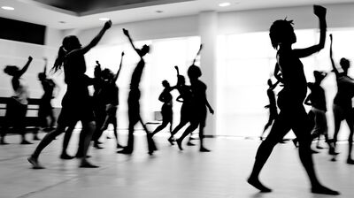 A black and white, motion-blurred photograph captures the silhouettes of a large group of dancers mid-movement in a bright dance studio.