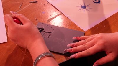 Hands with long nails use a tool to carve a butterfly design into a dark block on a wooden table.