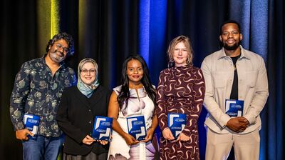Five smiling award winners stand in a row holding blue booklets against a black, yellow, and blue curtain backdrop.