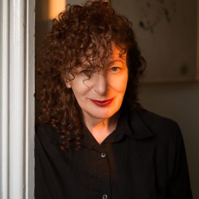 A woman with voluminous curly reddish-brown hair and red lipstick peeks from behind a white door frame into warm light.
