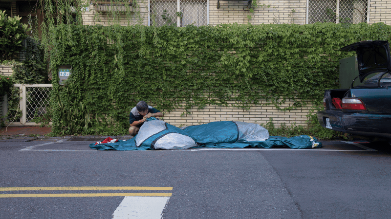 A man seemingly lifting the rear end of a large, teal inflatable replica car wedged onto a curbside parking space in front of a residential building.