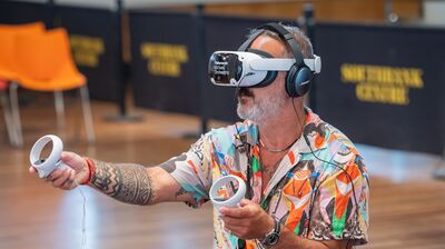 A man in a Hawaiian shirt wears a VR headset and headphones, holding two controllers in the clore ballroom