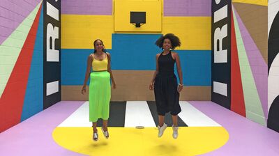 Two women jump in the air in front of a brightly colored, geometric mural on an outdoor basketball court.