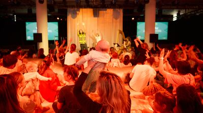 An audience of parents and babies watch a stage performance, with a mother holding her baby up in the foreground.