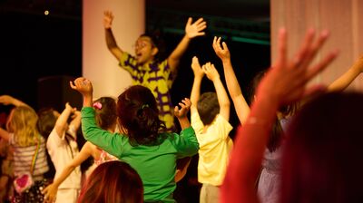 A group of children raise their hands while following along with a performer on a brightly lit stage.