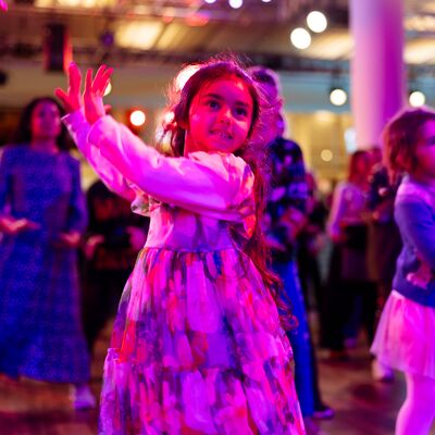 A young girl in a floral dress dances with other children under vibrant pink and purple lighting.