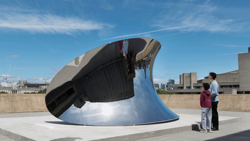 A gif of an outdoor wide shot shows two people standing on a concrete terrace looking at a large, curved, mirror-polished steel sculpture that reflects the sky and surrounding brutalist architecture and a silhouette of a person standing in front of a replica of the Earth