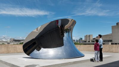 An outdoor wide shot shows two people standing on a concrete terrace looking at a large, curved, mirror-polished steel sculpture that reflects the sky and surrounding brutalist architecture.