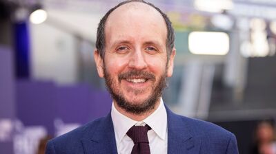 A close-up portrait of Jack Thorne smiling, wearing a blue suit, white shirt, and dark textured tie.