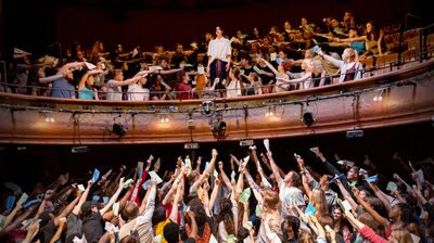 A woman in a white shirt stands in the centre of a theatre balcony, while the rest of the audience reach out towards here holding letters