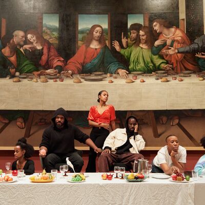 A group of seven people recreate Leonardo da Vinci's The Last Supper in front of a large reproduction of the painting.