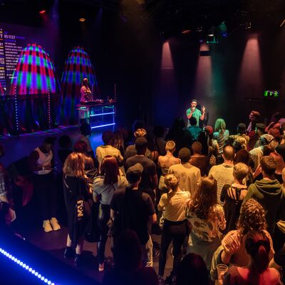An audience watching a poet with a DJ off to one side, in a studio theatre with multi-coloured lighting