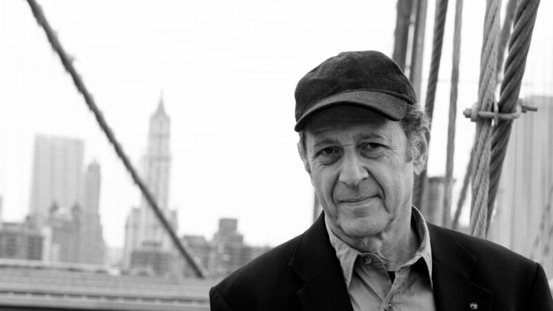 Steve Reich wearing a baseball cap and suit, standing on a bridge with the skyline of New York in the background