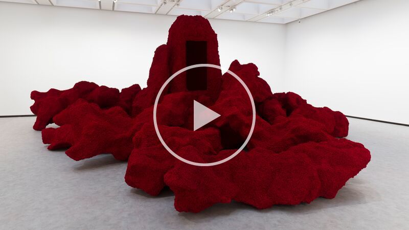 A large, textured dark-red sculpture resembling an organic rock formation sits in the center of a bright, white-walled museum gallery, featuring a tall central monolith with a void-like black rectangular opening.