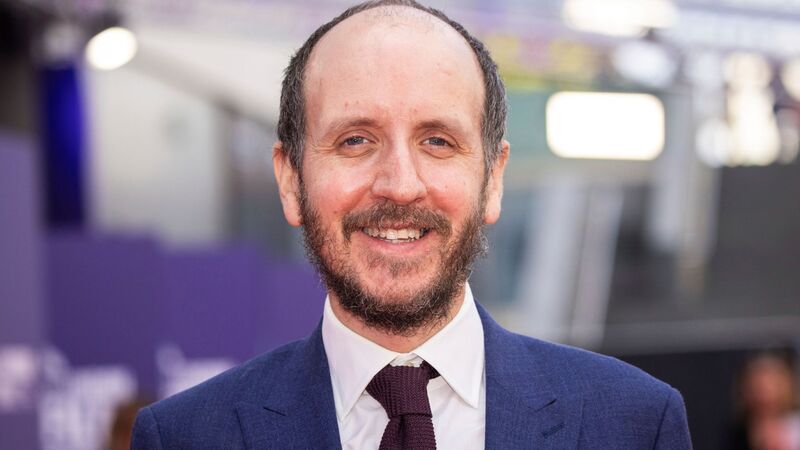 A close-up portrait of Jack Thorne smiling, wearing a blue suit, white shirt, and dark textured tie.