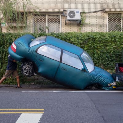 a man seemingly lifting the rear end of a large, teal inflatable replica car wedged onto a curbside parking space in front of a residential building.