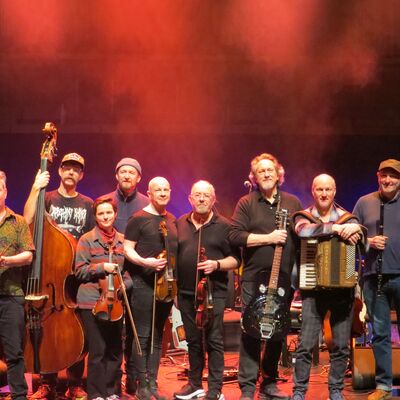 A group of musical performers line up on stage with various acoustic instruments under warm red lighting.