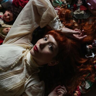 A redhead woman in a cream blouse lies among a collection of vintage porcelain dolls.