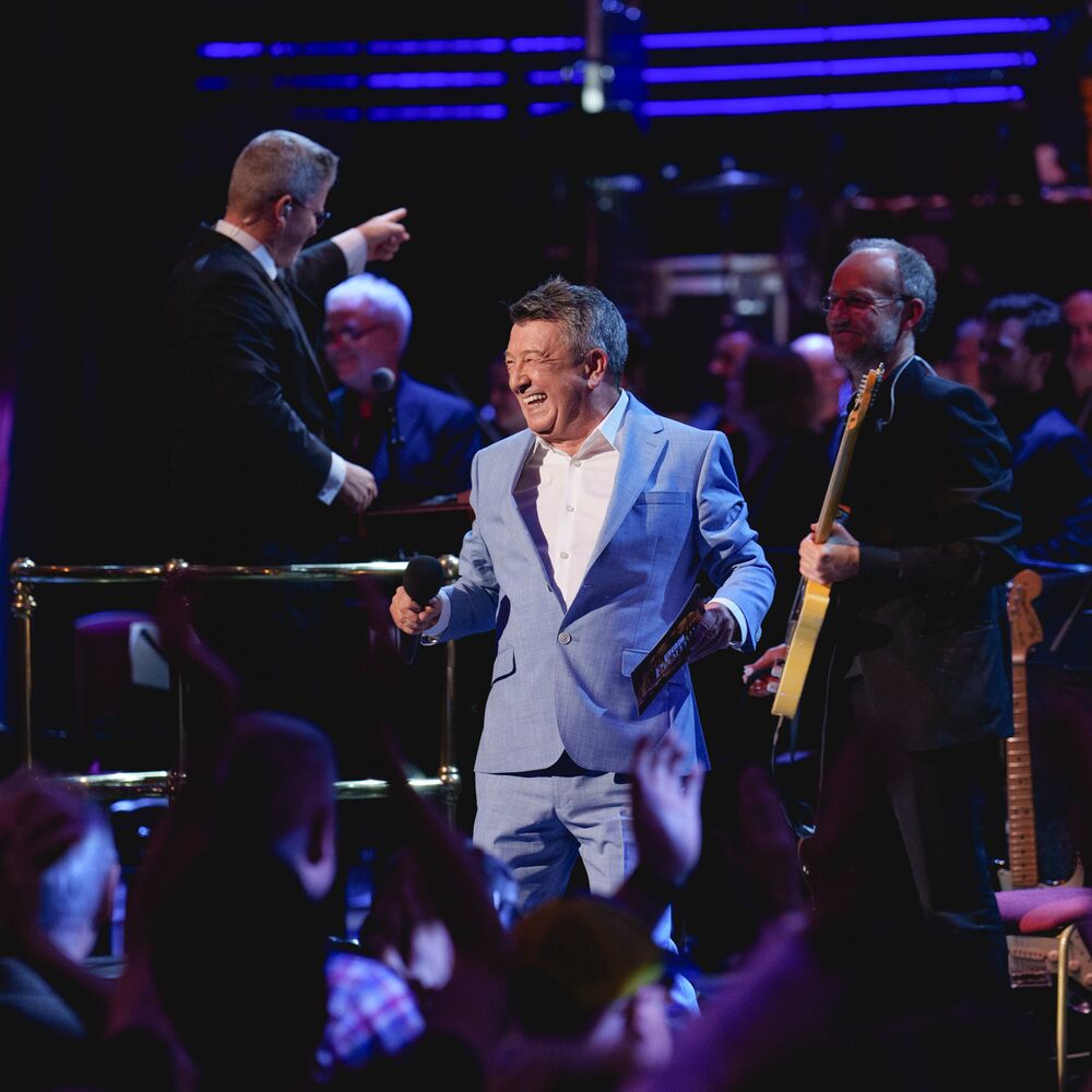 A performer in a light blue suit smiles on stage surrounded by musicians and an applauding audience.