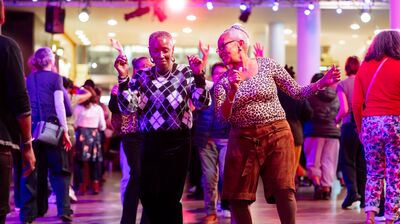 Two older women joyfully dance together in the center of a crowded room illuminated by vibrant pink stage lights.