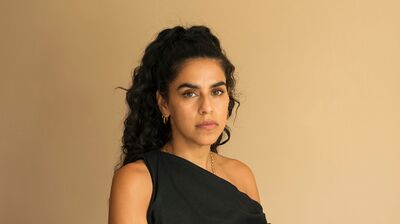 Woman with dark curly hair wearing a black dress slanted at the shoulder against a beige background.