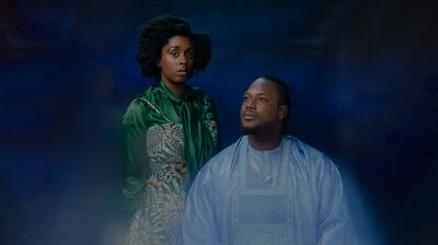 A woman in a green silk blouse stands behind a seated man in a light blue embroidered robe against a smoky dark blue backdrop.