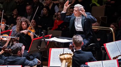 An elderly conductor in a tuxedo gestures expressively while leading an orchestra of violinists during a live concert.