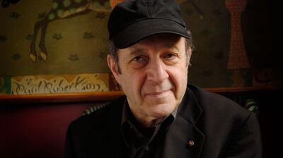 Composer Steve Reich in a black cap and jacket, posing in front of a dark tapestry featuring horses and riders.
