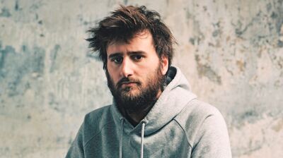 Portrait of a man with a messy beard and hair, wearing a gray hoodie against a weathered, neutral-toned wall.
