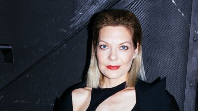 Close-up of a blonde woman with blue eyes and red lipstick posing in front of a textured black wall.