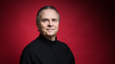 Portrait of a smiling man in a black high-collared shirt, posing against a solid deep red background.