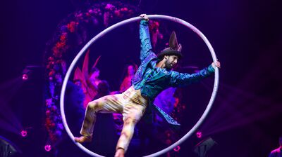 A performer in a green jacket and rabbit ears spins inside a large metal Cyr wheel under vibrant purple stage lights.