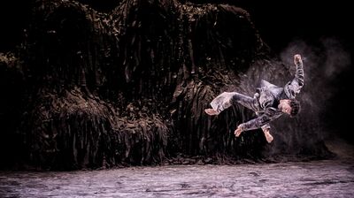 Performer wearing all black performing a flip through the air on a floury stage