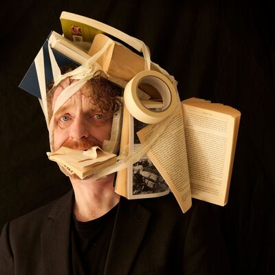 A man in a black blazer looking directly at the camera while a chaotic cluster of books and a roll of masking tape are bound tightly to his head and stuffed into his mouth using beige tape.