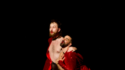 Two bearded performers in red sequined outfits and makeup pose expressively against a solid black background.